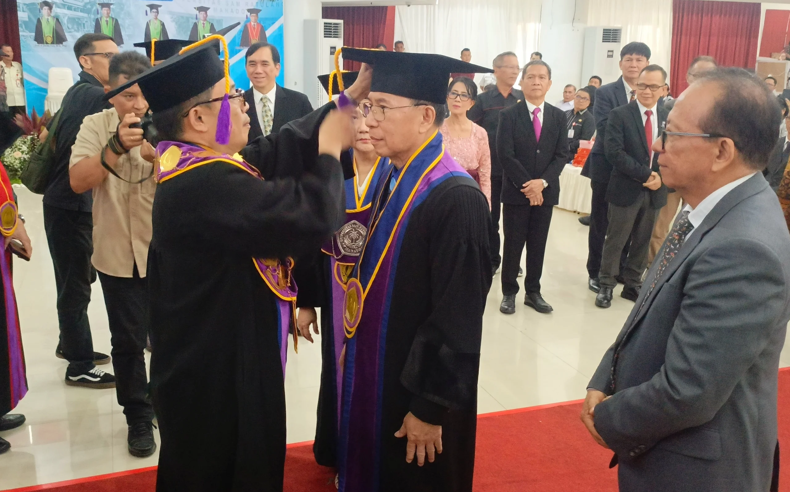 Rektor Berty Sompie saat melakukan prosesi pemakaian toga kepada Prof. Steenie Wallah dalam Sidang Senat Terbuka Universitas Sam Ratulangi. Momen ini menjadi puncak perjuangan akademik Prof. Steenie yang membawa inovasi Beton Geopolimer sebagai solusi konstruksi masa depan yang ramah lingkungan. (Foto: Ridho L Tobing)